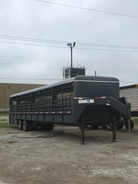 Black livestock trailer parked on gravel near a building under cloudy sky.